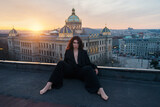 Fototapeta Zwierzęta - Attractive young curly brunette woman in a black jacket without lingerie sits on a rooftop against a historic building during a cozy sunset
