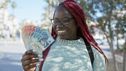 Canvas Print - A cheerful african woman with braids holding australian currency outdoors in the city.