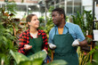 Experienced african american greenhouse owner and interested young female worker talking while standing in apron among green ornamental potted plants