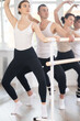 Young adult woman with group of fitness enthusiasts engaging in modern barre workout, performing demi plie with side lean, focusing on muscle strength and flexibility..