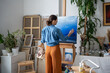 Professional female artist working on picture. Back view, woman concentratedly thoughtfully thoroughly drawing paintings using brush, oil paints, getting sense of peace calm from creative process.