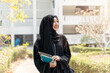 Closeup cheerful Muslim student wearing traditional clothes stand and holding textbook on walkway on her school building with sun flare background.