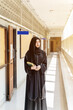 A Muslim student wearing traditional clothes standing and hold Quran on walkway in her school building with sun flare background.