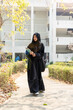 Asian Muslim wearing traditional clothes holding textbook and walking on walkway at her school building with sun flare background.