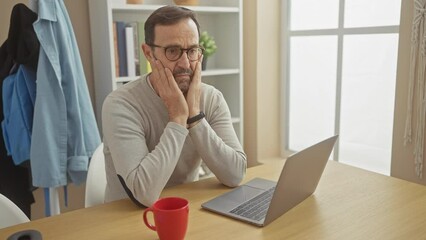 Poster - A pensive bearded middle-aged man works on a laptop at home, expressing focus and contemplation surrounded by calm domesticity.