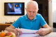 Serious older man writing letter at table in living room