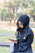 Young Asian beautiful woman Muslim medical student wearing traditional clothes relaxing with tablet in the hospital park with sun fare and blurred background.