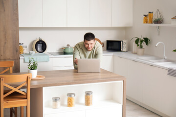 Wall Mural - Young bearded man using laptop on table in kitchen