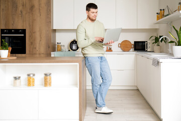 Wall Mural - Young bearded man using laptop in kitchen