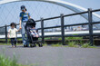 © kapinon - 晴天の川沿いの公園でベビーカーで散歩するママと兄弟の親子(3人家族)の全身　手を繋ぐ春夏のお出かけやママの通勤などのイメージ　