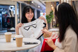 Two Asian women sit in a cafe at a mall, one excitedly showing off a new shirt to her friend.