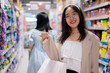 A smiling Asian woman stands in a grocery aisle, holding shopping bags, shopping in a supermarket.