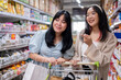 Two young Asian women shop together in a grocery store, focused on selecting items from the shelves.