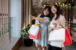 Two happy young Asian women pointing and smiling in a stylish shopping area, holding shopping bags.