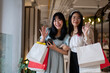 Two cheerful young Asian women wave happily at the camera while holding shopping bags.