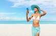 Happy young woman at the beach side using mobile phone, wearing a turquoise sun hat and bikini, portrait of African latin American woman in sunny summer day with blue sky, concept of a summer holiday