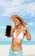 Happy young woman at the beach side showing mobile phone, wearing a turquoise sun hat and bikini, portrait of African latin American woman in sunny summer day with blue sky, concept of summer holiday