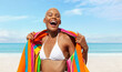 Happy woman at the beach side wrapped in a colorful towel, African latin American woman enjoying a sunny day with blue sky. Concept of summer beach holiday or booking travel and resort accommodations
