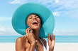 Happy woman at the beach side wearing blue sun hat and using mobile phone in a sunny day with blue sky. Concept of summer beach holiday, shopping online, booking travel and resort accommodations