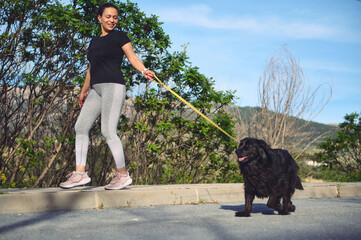 Wall Mural - Happy female athlete, woman runner walking her dog on leash with performing morning jog on the nature outdoors. People, nature, pets and healthy active lifestyle concept