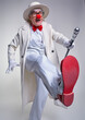 Clown in a white coat, hat and large red patent leather clown boots kicks forward with a joyful expression on his face.
