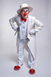 An elderly clown in a white coat and hat with walking stick with a cocky expression on his face and his hands on hips.