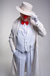An elderly serious mafia-like clown in a white coat and hat. Right hand in pocket, left hand adjusts the hat.