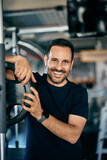 Fototapeta Pomosty - Portrait of a smiling male personal trainer, standing next to a squat rack, leaning on weights.