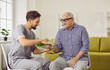 Portrait of a young smiling friendly physiotherapist, nurse or caregiver showing gray-haired senior man an exercise with a spiky ball for hand massage sitting at home in retirement.