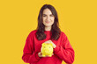 Finance. Portrait of smiling young woman who is holding piggy bank in form of pig on orange background. Caucasian woman in red sweatshirt is holding yellow piggy bank and looking at camera smiling.