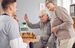 Young nurse man playing chess game with a happy smiling cheerful senior elderly couple sitting at the table in medical clinic. Caregiver support and retirement home leisure concept.