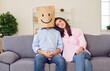 Love couple sits on sofa emotions man in paper bag with drawn smile woman smiling emotions of joy. Charming emotions illustrates warm and happiness in this family Man in paper bag with smile