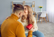 Happy young couple with red gift box hugging at home. Attractive romantic man and woman wearing knitted pullovers embracing, kissing with tenderness. Family couple celebrating event with present box