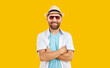 Portrait of a confident happy smiling young bearded handsome man standing with crossed arms wearing casual summer clothes in hat and sunglasses isolated on studio yellow background.