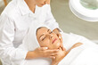 Young woman with closed eyes and with white towel on head lying under magnifying beauty lamp enjoying professional face and neck massage done by her beautician. Skin care, facial treatment concept