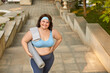 High angle shot of happy cheerful smiling young fat plump woman in fitness activewear, with towel on shoulder, standing on stone steps in city park, holding workout mat, looking at camera and smiling