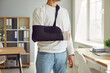 Close-up cropped photo of a man's broken arm in a plaster cast standing at home or at office on rehabilitation after injury. Patient wearing sling or bandage. Recovery and rehab concept