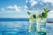 Two cocktail glasses decorated with lime and mint on an infinity pool that merges with the sea, on a background of blue sky with white clouds
