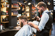 Professional barber using comb and electric shaver to cut side hair. Male customer getting stylish haircut. Side view of man on background of other hairdresser.