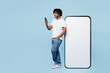 © ViDi Studio - Full body side view young fun Indian man wear white t-shirt casual clothes big huge blank screen mobile cell phone with area use smartphone isolated on plain pastel blue background. Lifestyle concept.