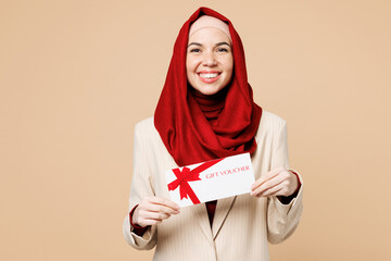 Wall Mural - Young Arabian Asian Muslim woman wears red abaya hijab suit clothes hold gift certificate coupon voucher card for store isolated on plain beige background. UAE middle eastern Islam religious concept.