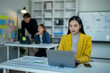 © Wasana - A woman in a yellow jacket is working on a laptop in front of a white board. She is surrounded by other people in the room, some of whom are looking at her. The atmosphere seems to be focused