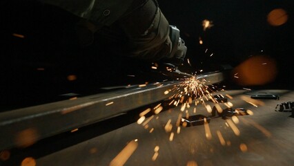 Craftsman in protective gloves working with grinder at industrial plant, man grinding iron detail, sparks flying at the camera.