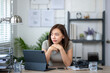 © Wasana - A woman is sitting at a desk with a laptop and a tablet. She is looking at the laptop screen and she is deep in thought