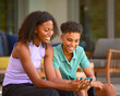 © Monkey Business - Teenage Brother And Sister Looking At Mobile Phone Sitting Outdoors At Home On Deck