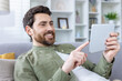 A cheerful, bearded man comfortably using a digital tablet while sitting on a couch. His relaxed posture and smile suggest he is enjoying his activity.