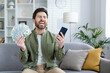 Exuberant male in a casual outfit joyously celebrating with a stack of cash and a smartphone in a cozy living room setting.