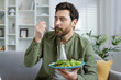 A bearded man eating a healthy green salad at home, seated comfortably in a stylishly furnished living room surrounded by plants and natural light.