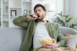 A bored young man yawning while holding a TV remote and a bowl of popcorn, sitting on a sofa in a living room setting.