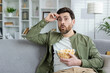 A man sits on a couch, looking shocked while watching a movie. He is casually dressed and holding a bowl of popcorn, expressing surprise.
